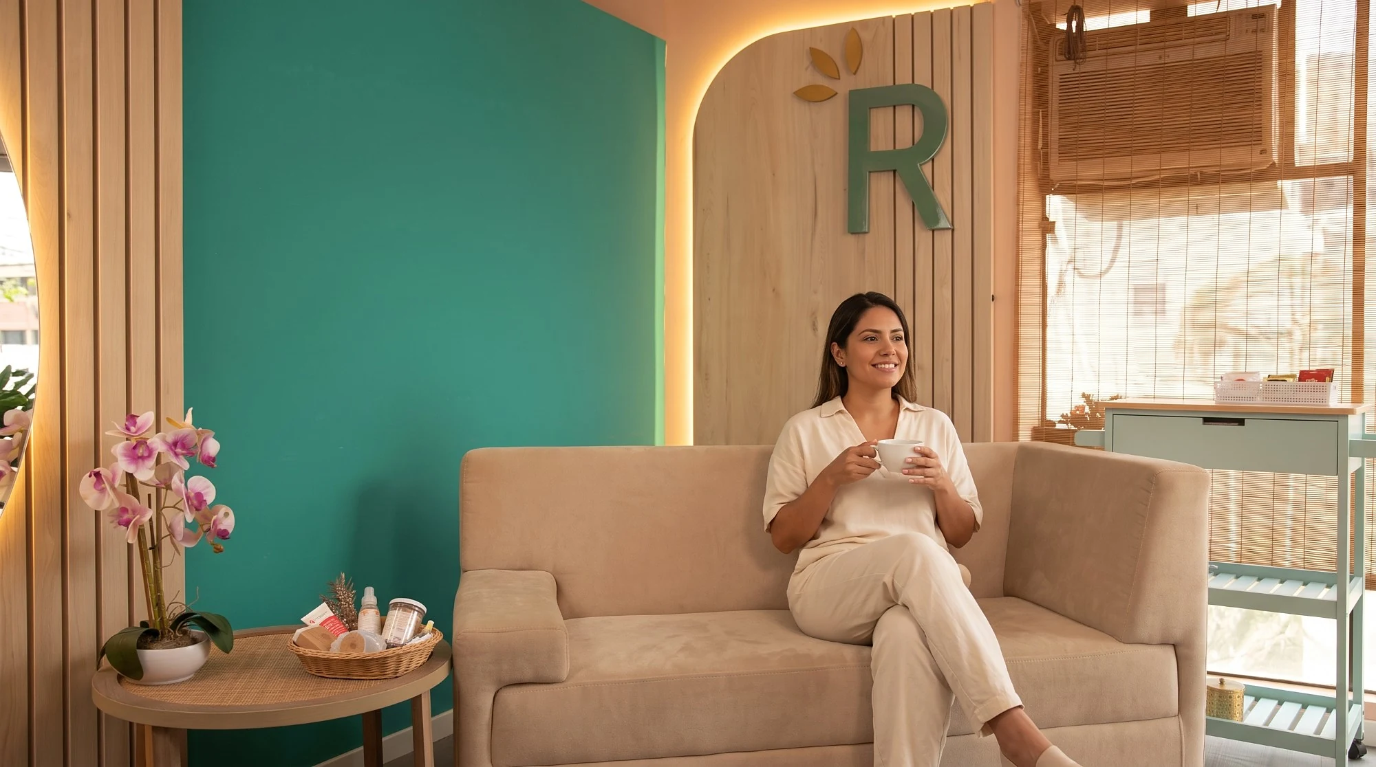Clienta sonriente con una taza de té en la sala de espera de Radiance, junto a la pared verde y la R del logo